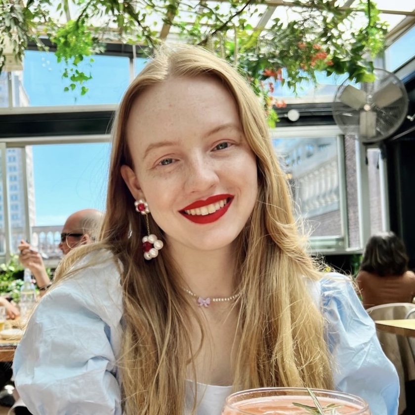 A picture of Domino Weir. She has strawberry blonde hair and blue eyes. Her skin is pale with lots of freckles. She is wearing red lipstick and a blue dress accessorized with pearl jewelry. She is smiling and framed by a view of a rooftop restaurant.